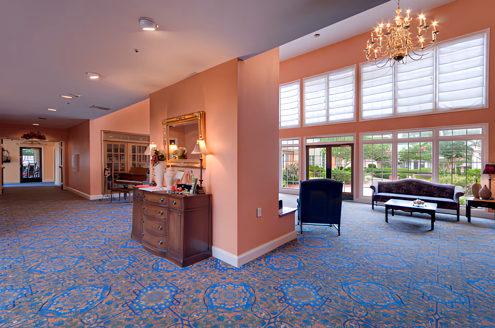 Bright, spacious lobby-style sitting area with upholstered chairs and sofas, a chandelier, and large windows overlooking the grounds.
