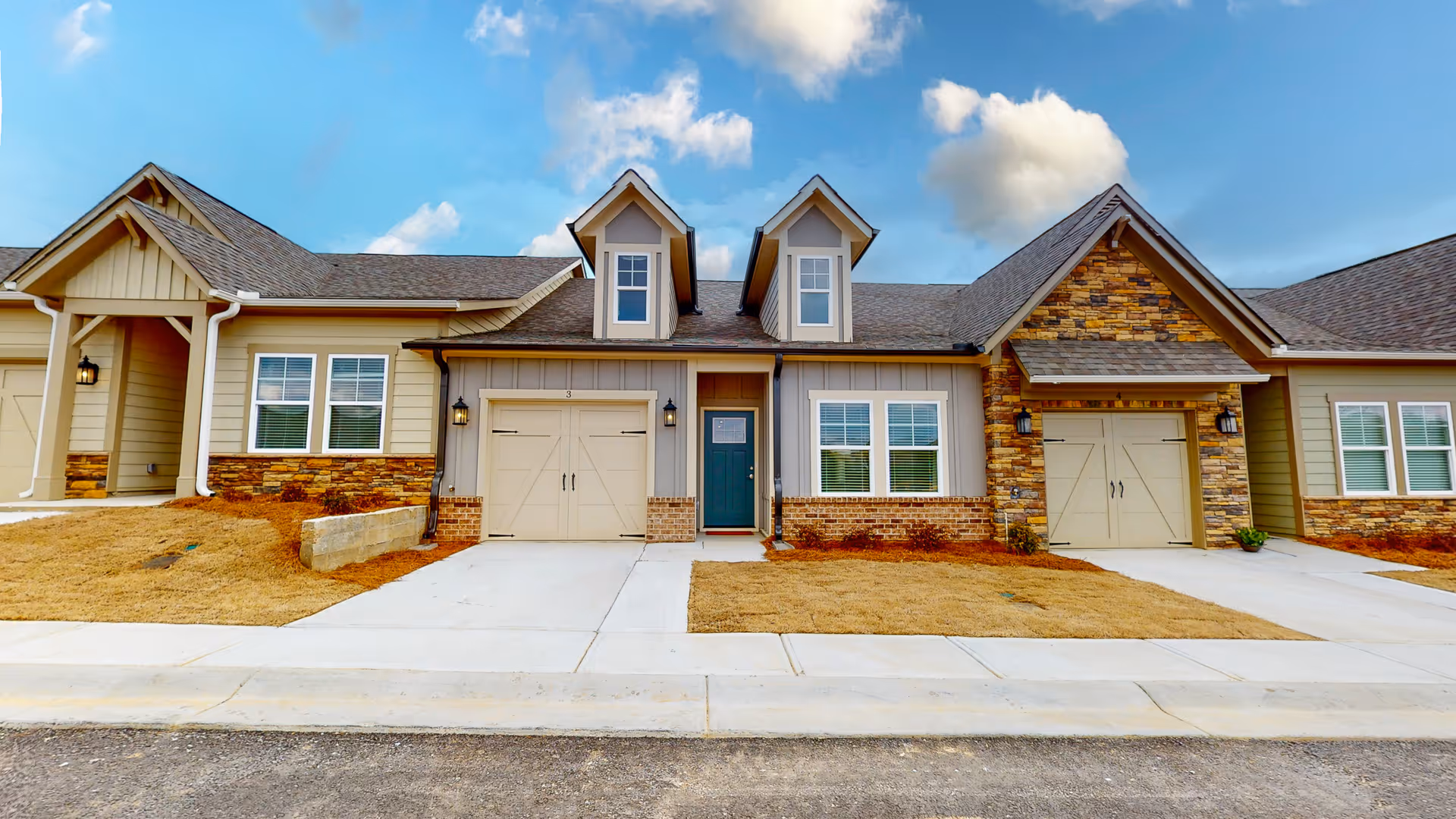 Exterior view of a single-story senior living facility building with beige and stone facade, two dormer windows on the roof, two garage doors, a central entrance door, and a concrete driveway under a partly cloudy sky.