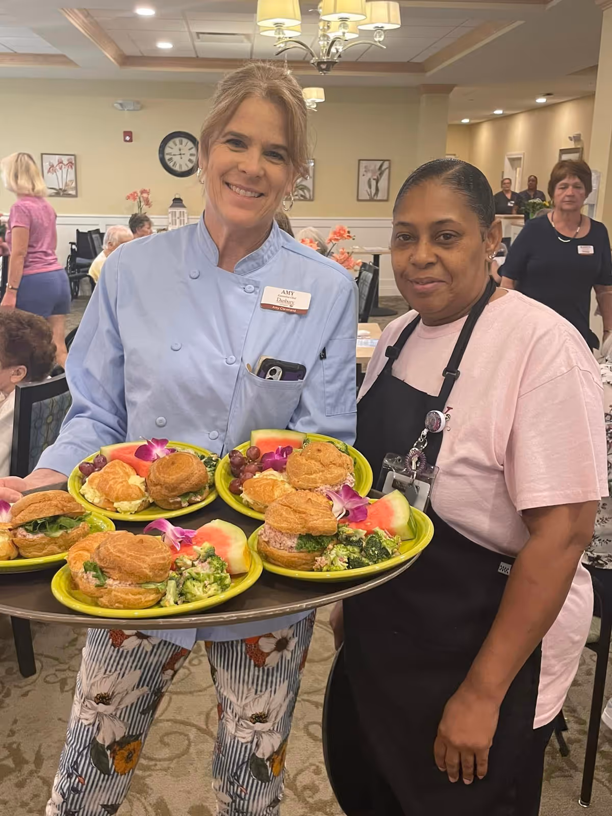 Two women standing in a dining area of a senior living facility. One woman, wearing a light blue chef coat and floral pants, is holding a tray with five plates of food, each plate containing a sandwich, broccoli salad, grapes, and a slice of watermelon garnished with a purple flower. The other woman, wearing a pink shirt and black apron, stands beside her. In the background, several elderly residents and staff members are visible in a well-lit room with beige walls and decorative wall art.