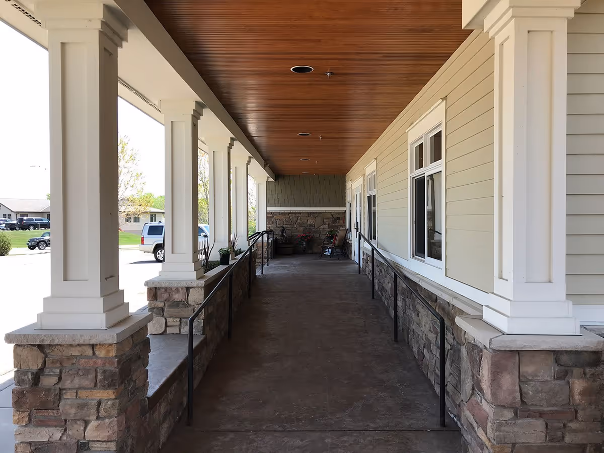 Covered outdoor walkway with stone pillars and white columns, a wooden ceiling, and a concrete floor with handrails on both sides. There are windows on the right wall and some chairs and plants at the far end of the walkway.
