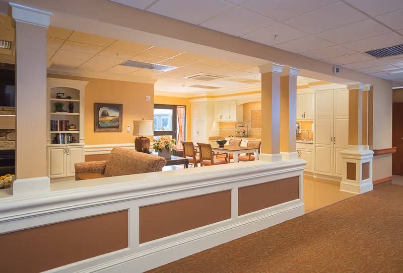 Interior view of a senior living facility showing a cozy living room area with a sofa, armchair, lamp, and bookshelf, adjacent to a kitchen and dining area with a table and chairs. The space is warmly lit with beige walls and white cabinetry.