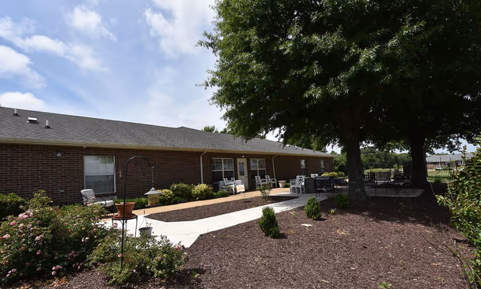 Outdoor patio area at Dogwood Pointe Senior Living featuring a paved walkway, garden beds with shrubs and flowers, several seating areas with chairs and tables, and large trees providing shade under a partly cloudy sky.