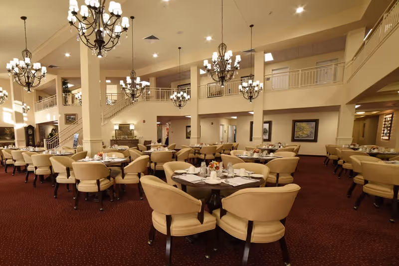 A spacious dining room with multiple round tables set with white dishes, cups, and silverware. The room features beige upholstered chairs, red carpet flooring, cream-colored walls, and several ornate chandeliers hanging from the ceiling. There is a staircase leading to an upper level with a railing, and framed artwork on the walls.