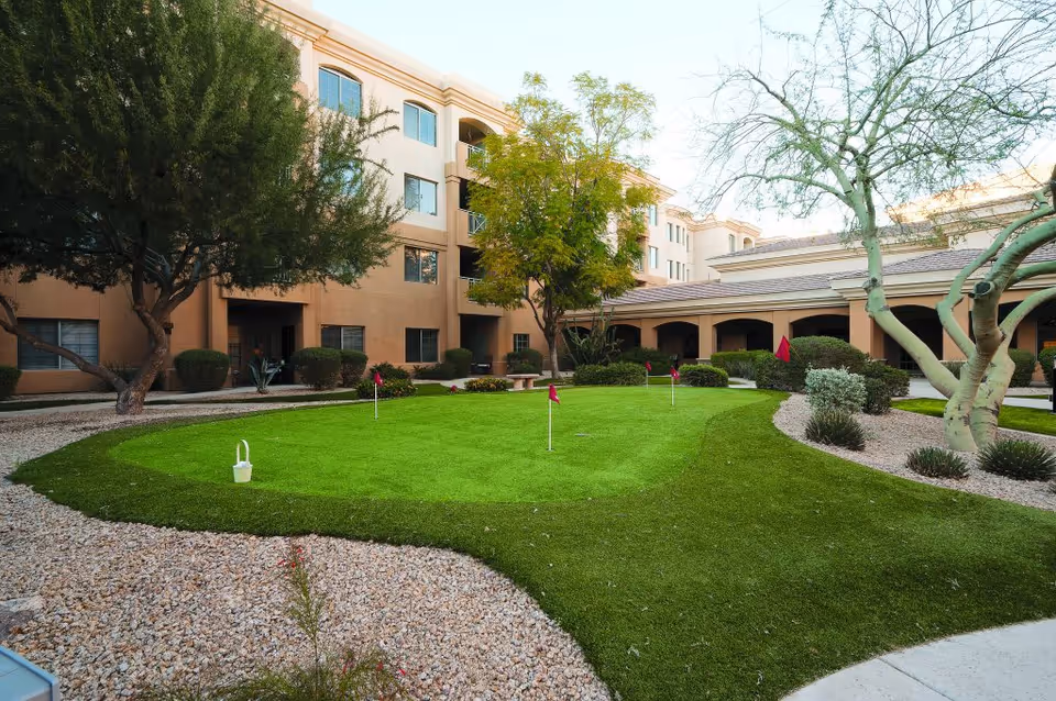Outdoor courtyard area of a senior living facility featuring a small putting green with several red flags, surrounded by gravel landscaping, trees, and bushes. The courtyard is enclosed by a multi-story beige building with windows and covered walkways.