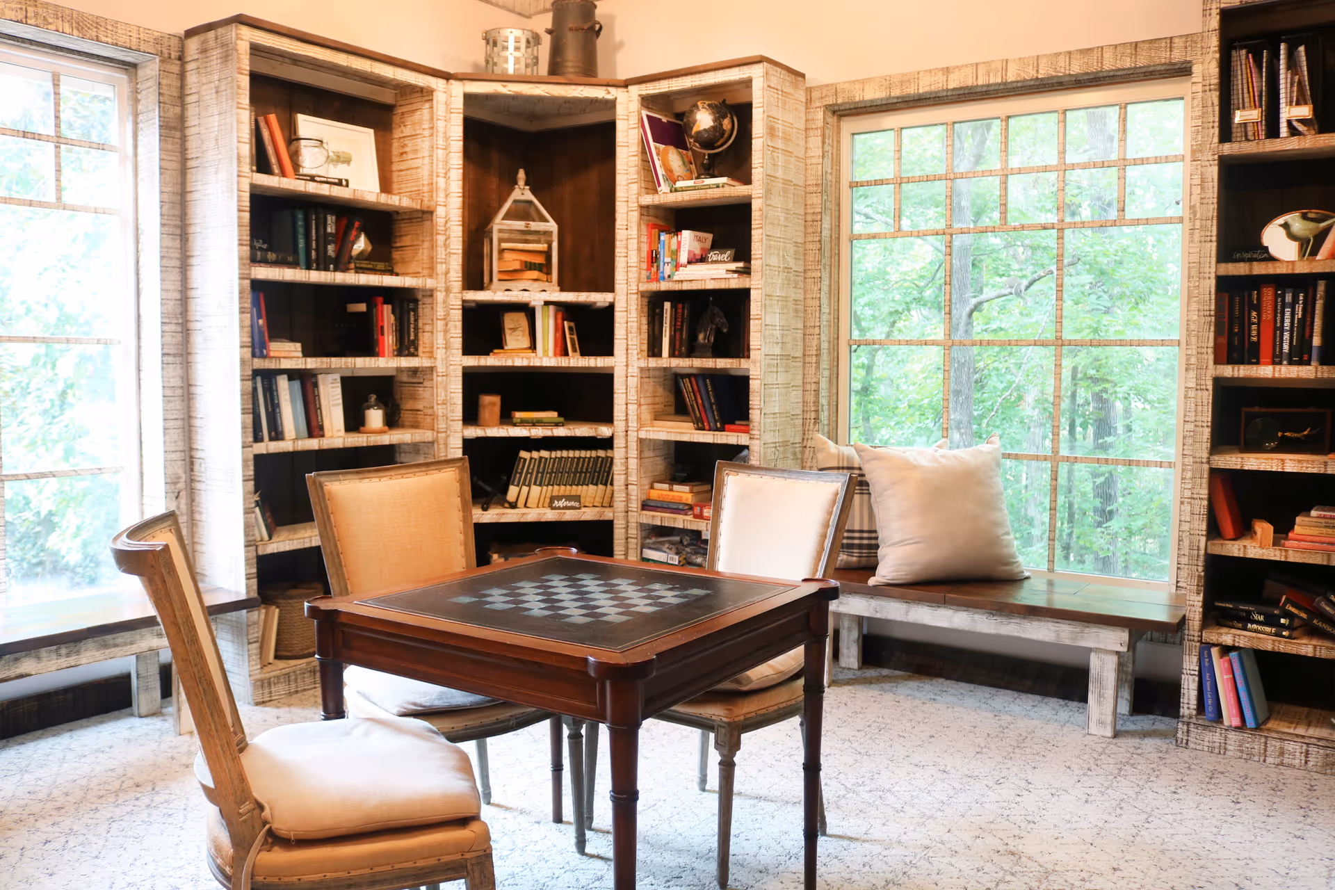 A cozy reading or game room with a wooden chess table and two cushioned chairs. Surrounding the table are built-in rustic wooden bookshelves filled with books and decorative items. Large windows let in natural light and offer a view of green trees outside. A bench with pillows is positioned under one of the windows.