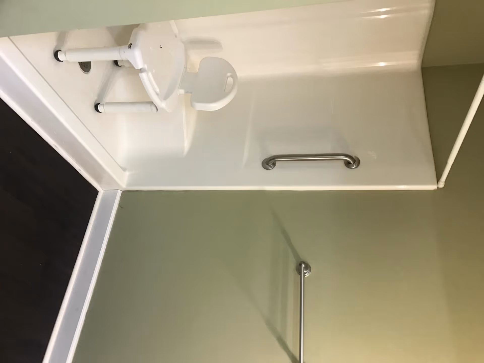A bathroom shower area with a white plastic shower chair and a metal grab bar mounted on the wall. The walls are light green and the floor is dark wood or laminate.
