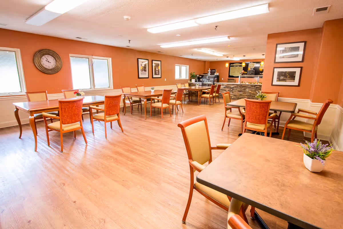 A spacious dining room with several wooden tables and chairs arranged neatly. The walls are painted a warm orange color, decorated with framed pictures and a large round clock. The room has large windows letting in natural light and a serving counter with a menu board in the background.