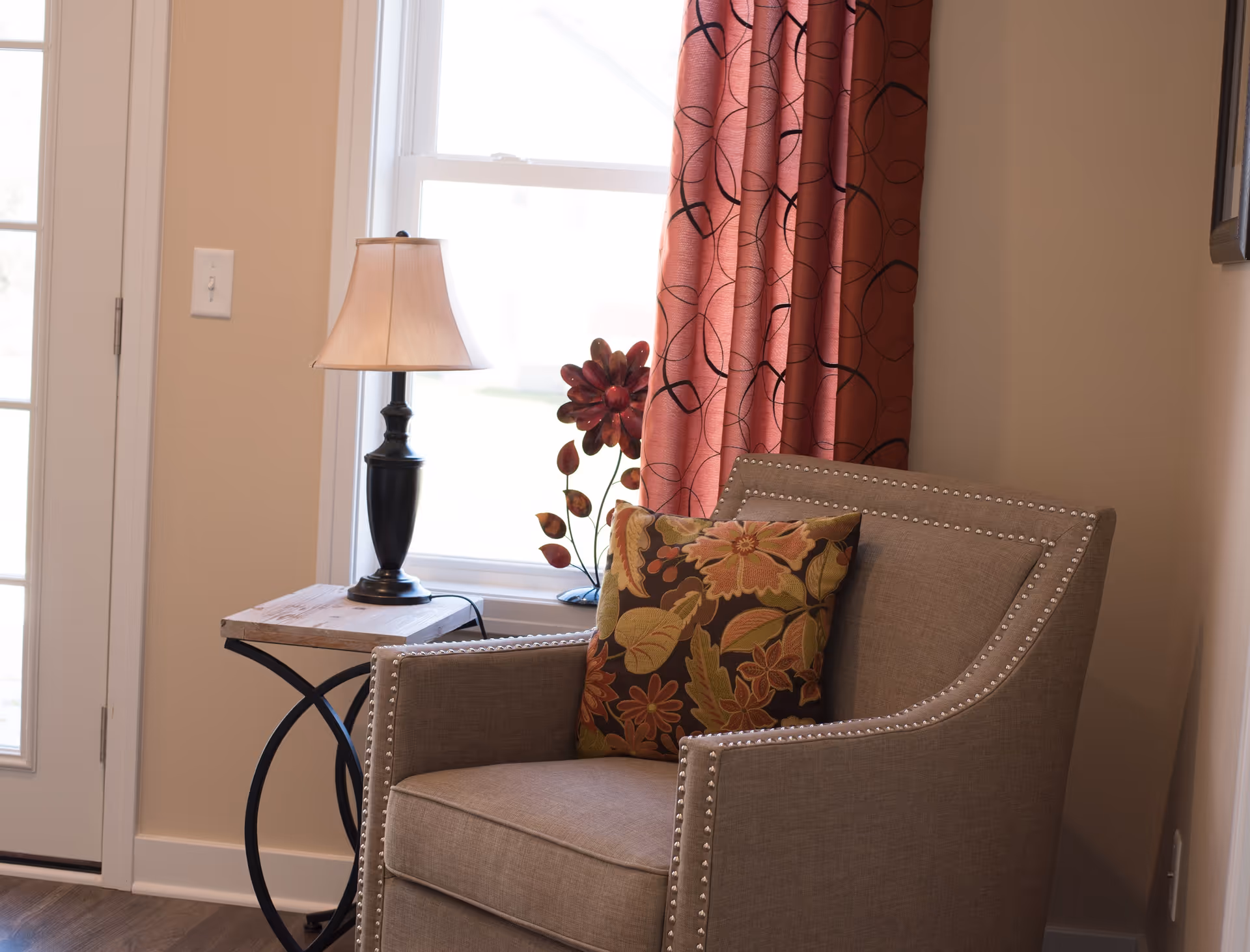 A cozy corner of a living room featuring a beige armchair with nailhead trim and a floral patterned cushion. Next to the chair is a small wooden side table with a black lamp and a decorative flower sculpture. Behind the chair, there is a window with red curtains that have a black swirling pattern.