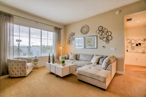 A bright living room with a large window covered by sheer curtains, a beige sectional sofa with cushions, a tufted armchair, and a modern coffee table with decorative items. The walls are adorned with circular metal wall art and framed pictures. The carpet is light brown, and there is a hallway visible to the right.