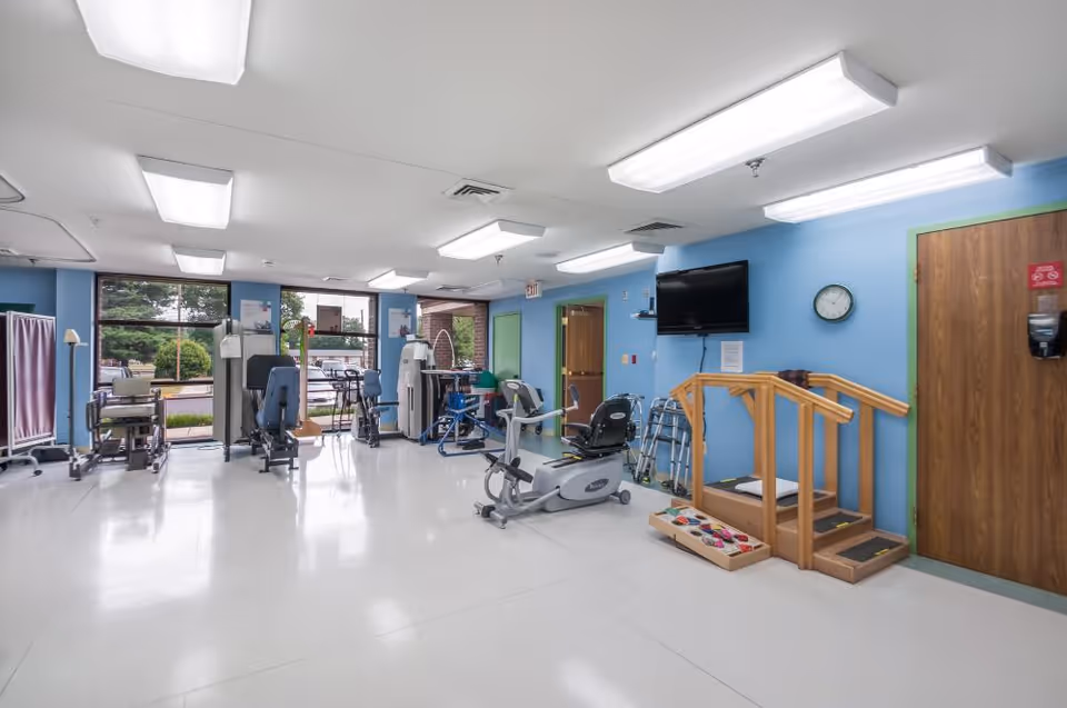 A spacious physical therapy or rehabilitation room with exercise equipment including stationary bikes, a leg press machine, parallel bars for walking practice, walkers, and a TV mounted on a blue wall. The room has large windows letting in natural light and a door with a green frame.