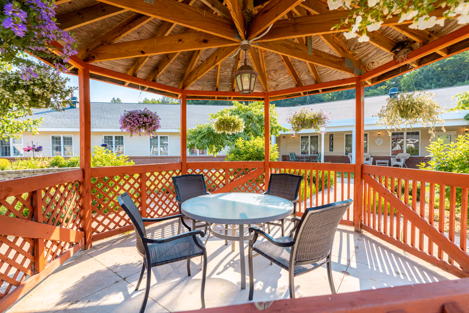 A wooden gazebo with a round glass table and four black mesh chairs inside. The gazebo is surrounded by hanging flower baskets and overlooks a courtyard with greenery and a single-story building in the background.