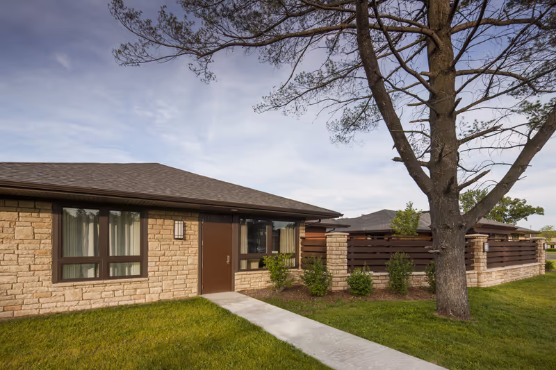 Exterior view of a single-story building with stone walls and a brown door, surrounded by green grass and a tree in the foreground under a partly cloudy sky.