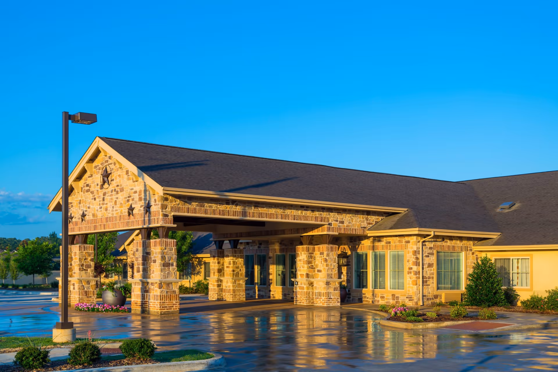 Exterior view of a single-story building with stone and brick facade, featuring a covered entrance supported by stone pillars. The building has a dark shingled roof and several windows with blinds. There are landscaped bushes and flowers around the entrance, and a street lamp is visible on the left side. The ground appears wet, reflecting the building and the clear blue sky above.