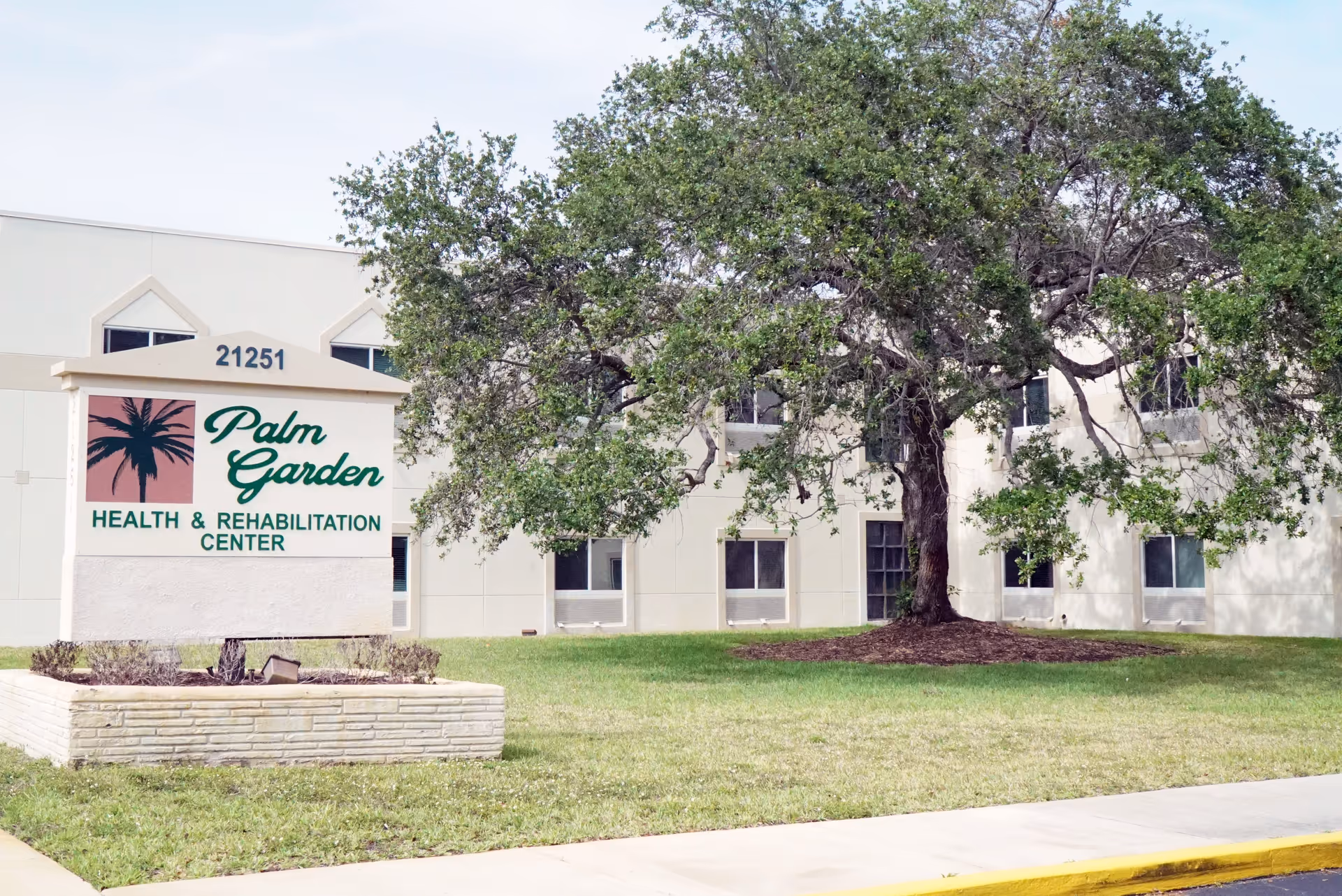 Exterior view of Palm Garden Health & Rehabilitation Center building with a large tree and green lawn in front. The building is light-colored with several windows, and a sign displaying the facility name and address 21251 is visible.