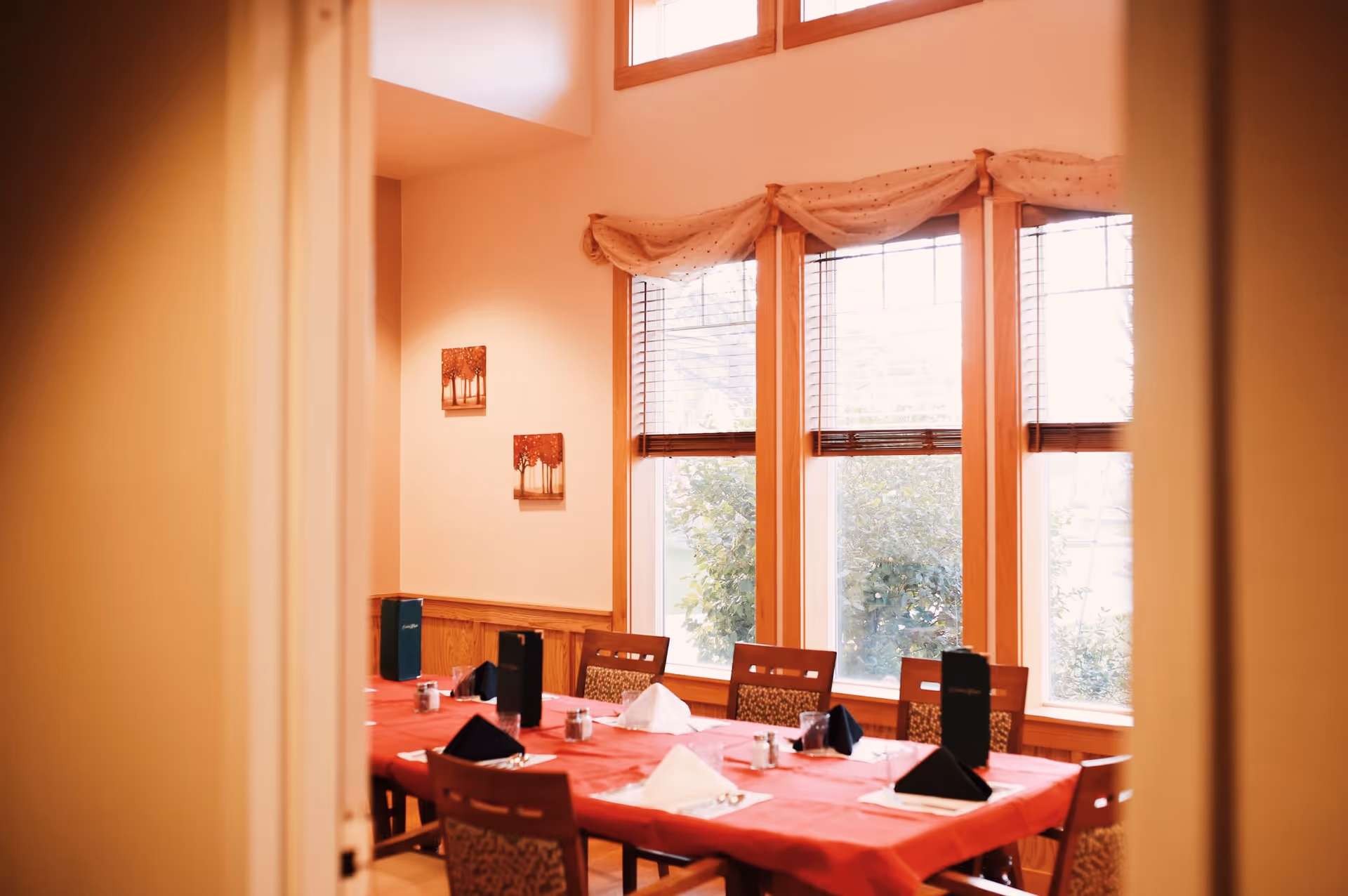 Dining room with a long table covered with a red tablecloth, set with folded napkins, menus, and salt and pepper shakers. The room has large windows with wooden frames and blinds, and two small paintings on the wall.