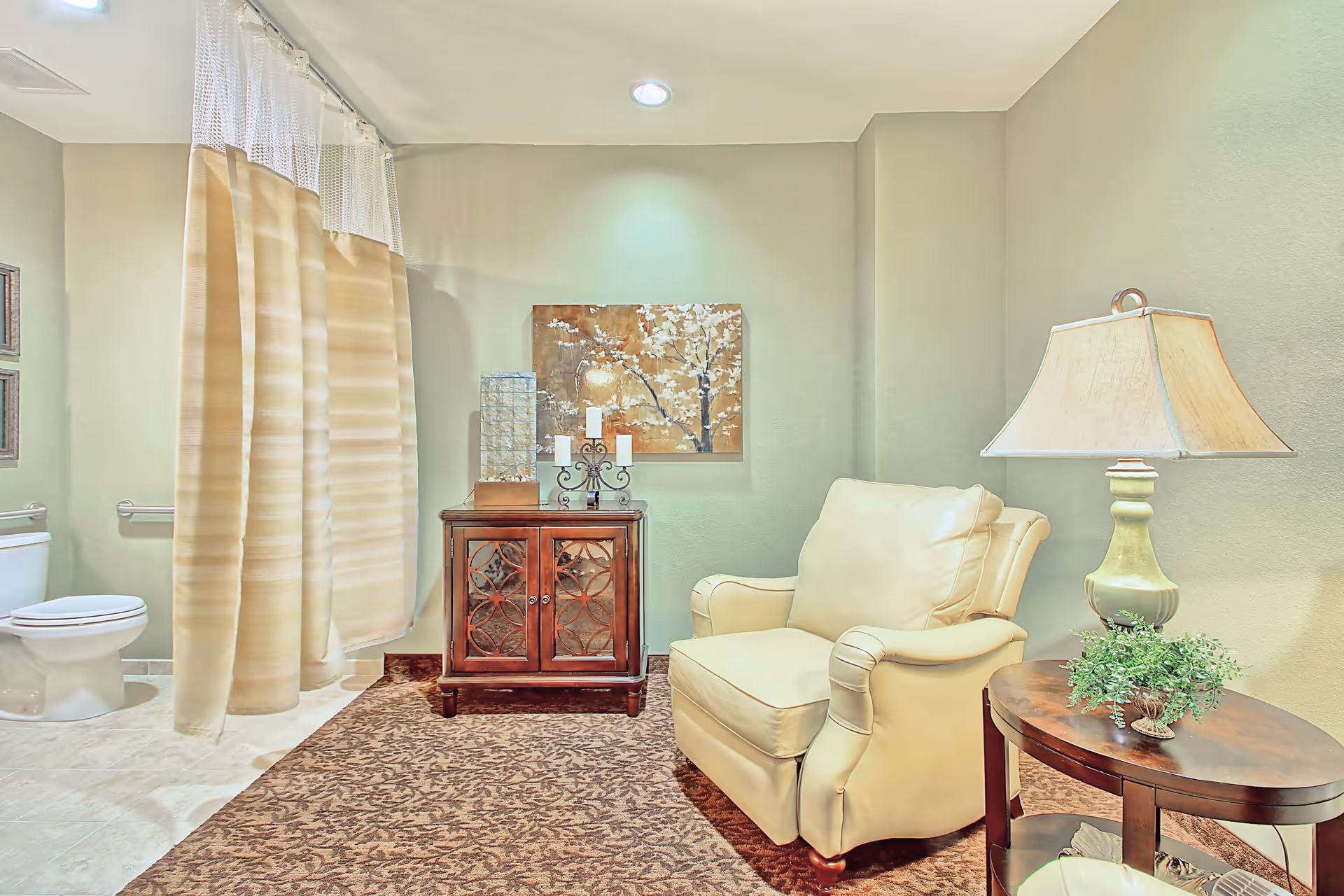 Cozy sitting area with a leather armchair, side table and lamp beside a decorative cabinet, with a toilet and shower curtain visible to the left.