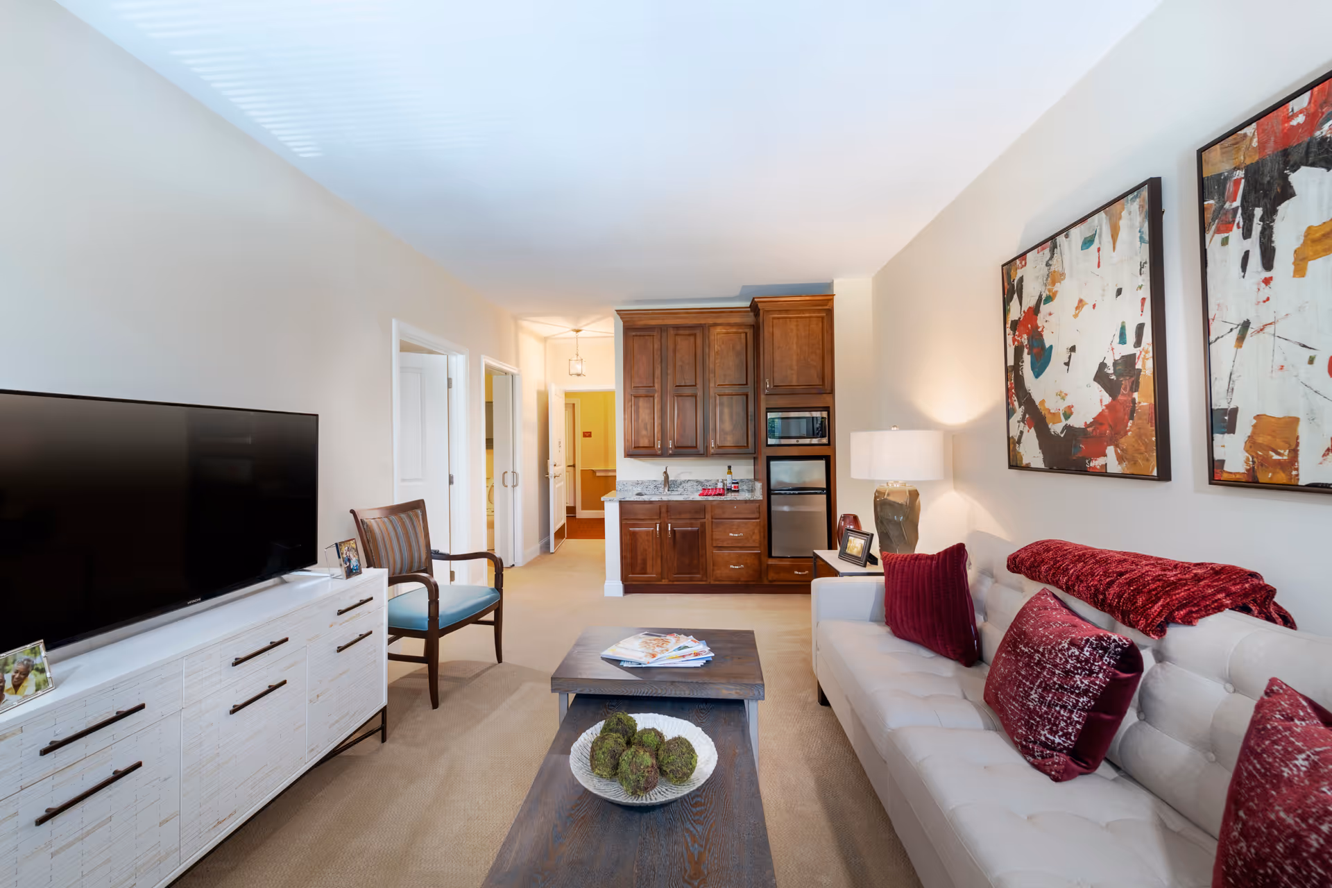 A bright and cozy living room in a senior living facility featuring a beige sofa with red and burgundy pillows and a matching throw blanket. A wooden coffee table with decorative items sits in front of the sofa. On the left side, there is a white media console with a large flat-screen TV and a wooden chair with a blue cushion. The back wall has a small kitchenette with wooden cabinets, a microwave, and a mini refrigerator. Two abstract paintings hang on the wall above the sofa, and a table lamp provides warm lighting.