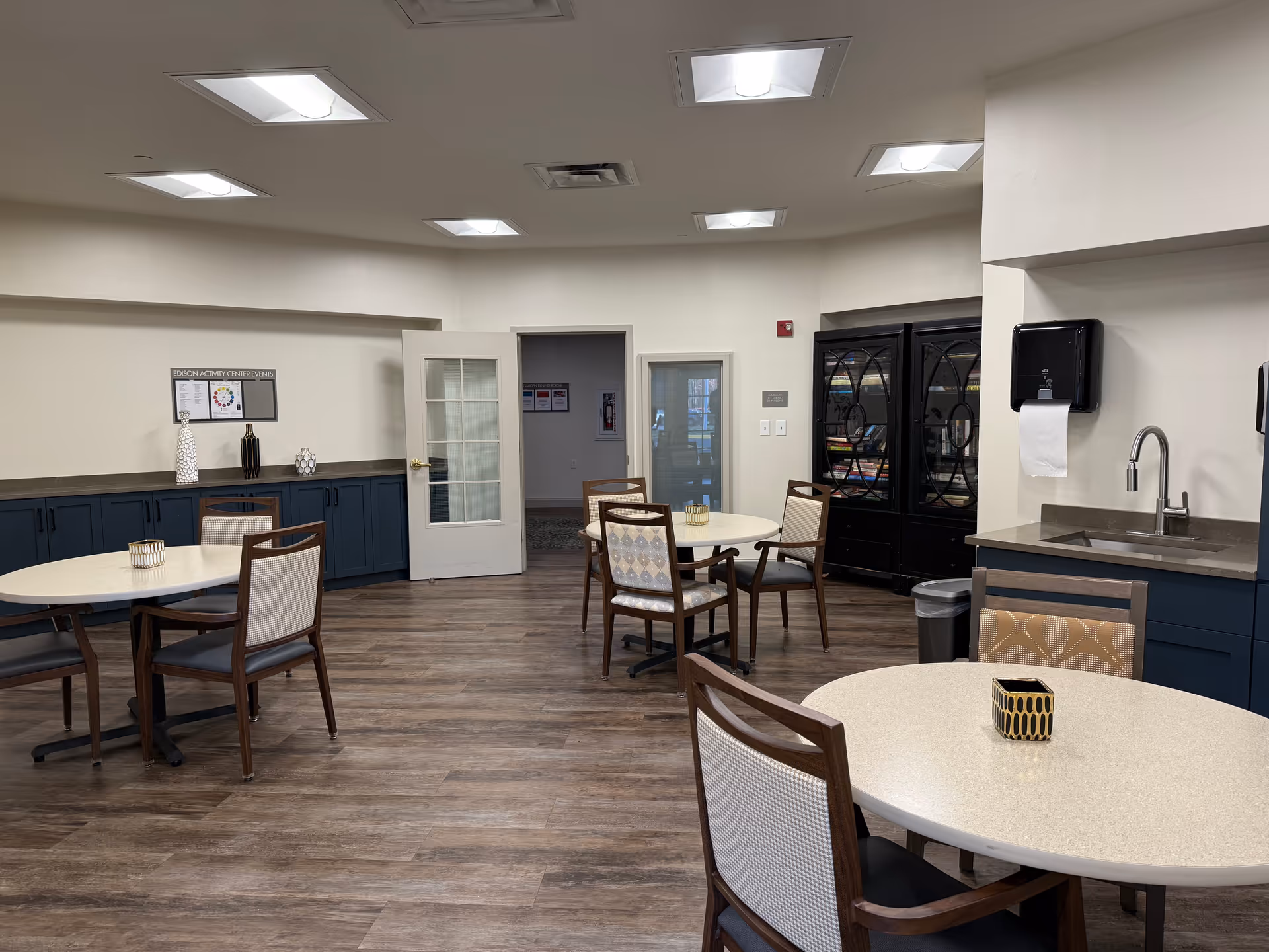 Interior view of a senior living facility activity room with round tables and chairs arranged on a wood floor. There are cabinets along the walls, a sink with a paper towel dispenser, and a glass-fronted cabinet filled with books. The room is well-lit with ceiling lights and has a door leading to another room.
