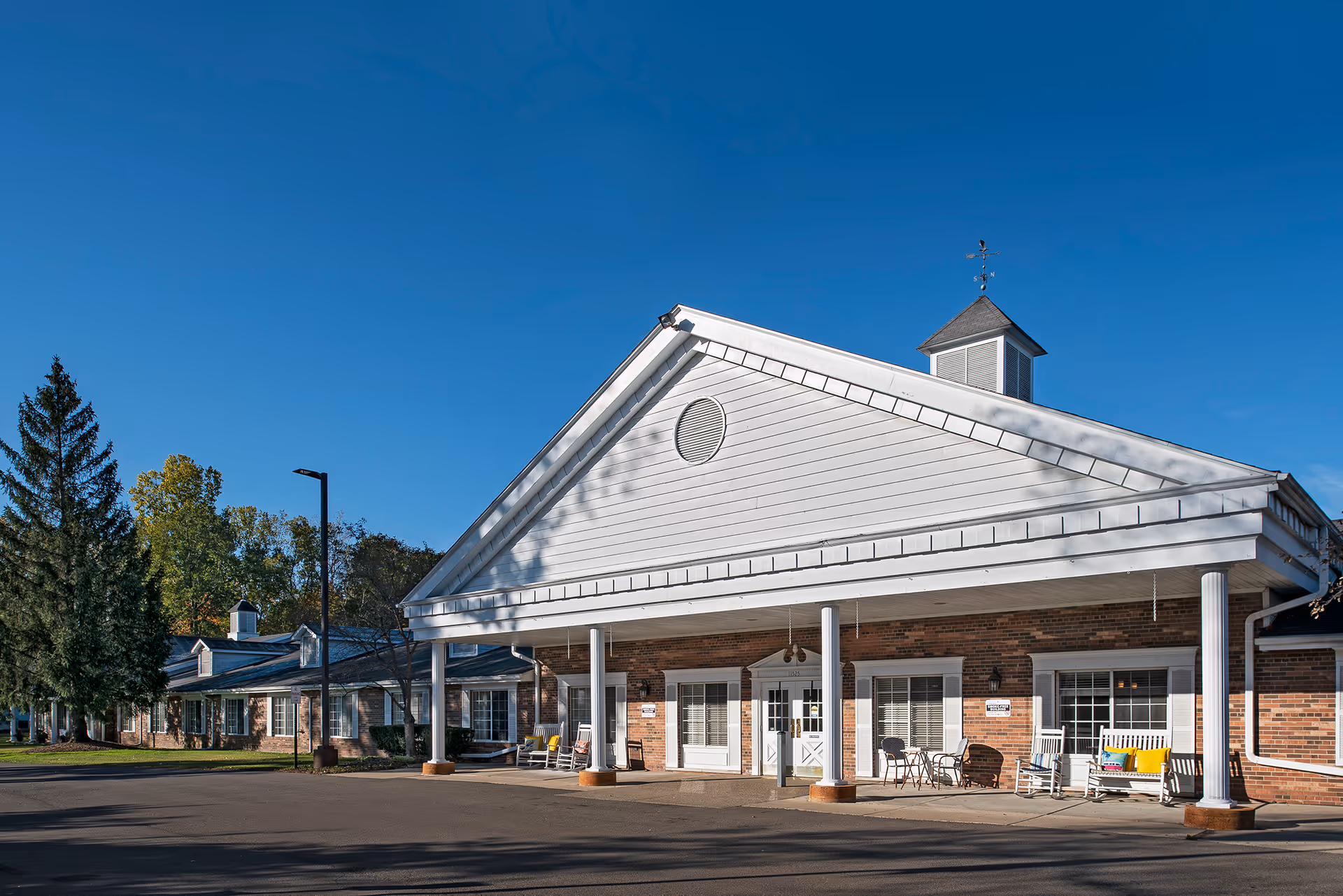Exterior view of a single-story senior living facility building with a large white gabled entrance supported by columns. The building has brick walls and white trim, with rocking chairs and a small table on the front porch. Trees and a clear blue sky are visible in the background.
