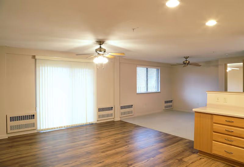 An empty living space in a senior living facility with wood flooring in the foreground and carpeted area in the background. The room features two ceiling fans with lights, a large window with vertical blinds, a smaller window with horizontal blinds, and a kitchen counter with wooden cabinets on the right side.