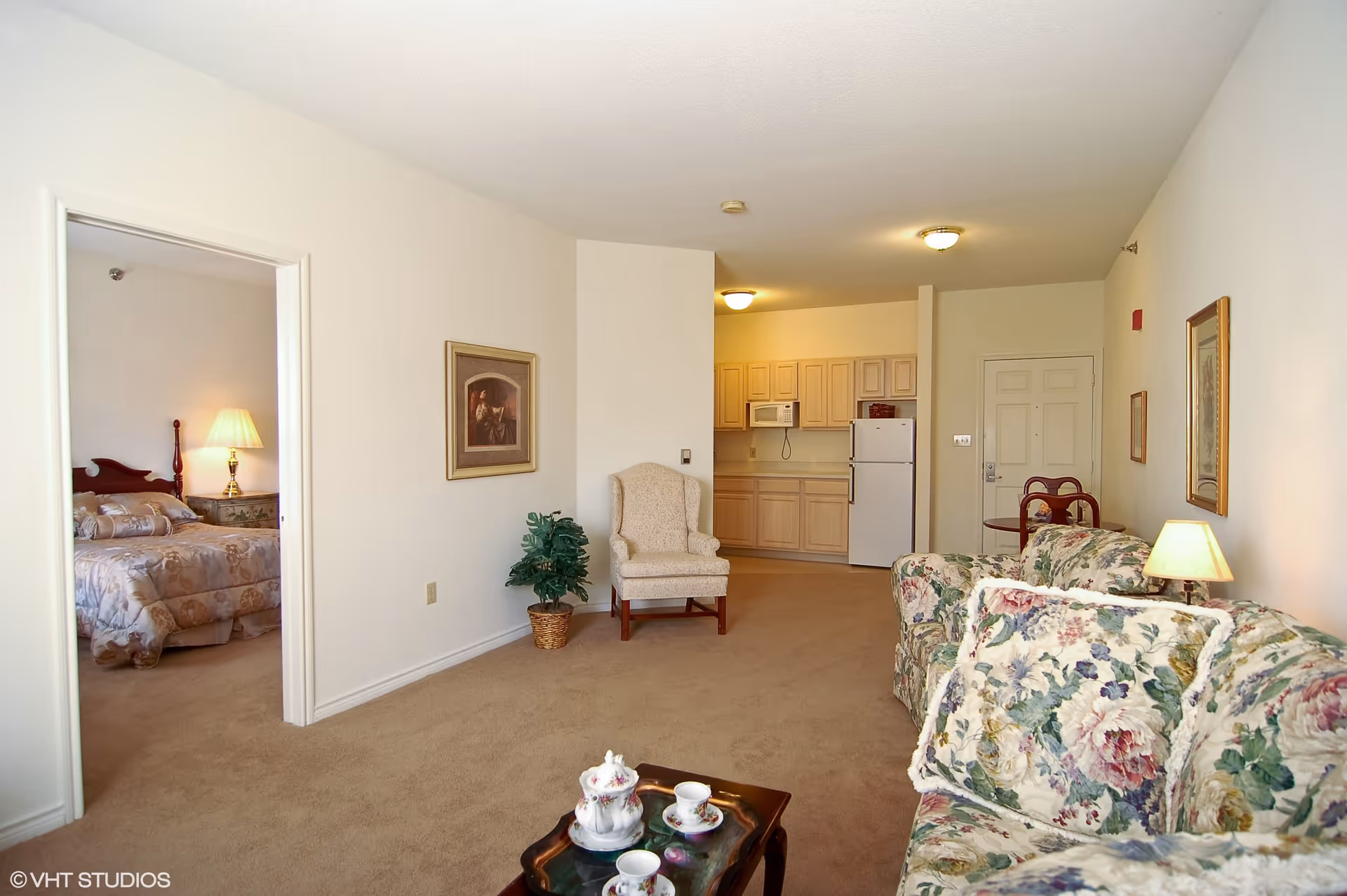Interior view of a senior living apartment showing a living room with floral patterned couches, a small coffee table with a tea set, a beige armchair, and a kitchen area with light wood cabinets, a microwave, and a refrigerator. A bedroom with a bed and a lamp is visible through an open doorway.