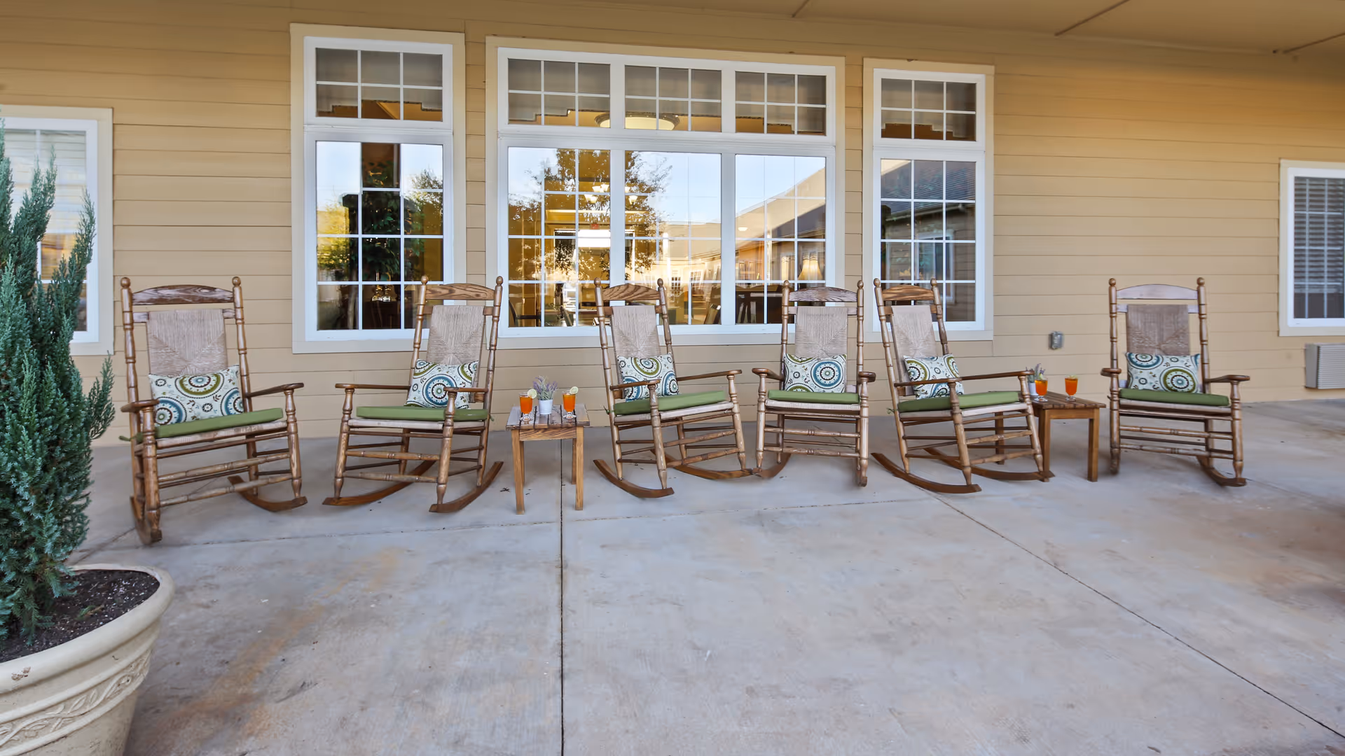 A row of six wooden rocking chairs with green cushions and patterned pillows arranged on a concrete patio outside a building with beige siding and large white-framed windows. Two small wooden tables between the chairs hold glasses with orange and clear beverages. A large potted plant is visible on the left side.