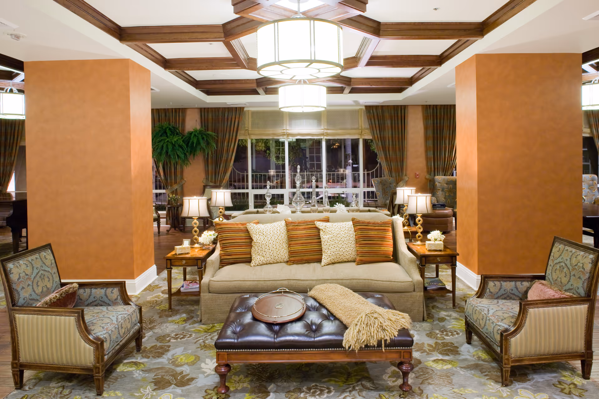 A cozy living room area with a beige sofa adorned with striped and patterned cushions, flanked by two wooden armchairs with patterned upholstery. In front of the sofa is a dark leather tufted ottoman with a tray and a beige throw blanket. The room features warm orange walls, a decorative ceiling with wooden beams, and large windows with curtains in the background. Two side tables with lamps and small floral arrangements are placed beside the sofa.