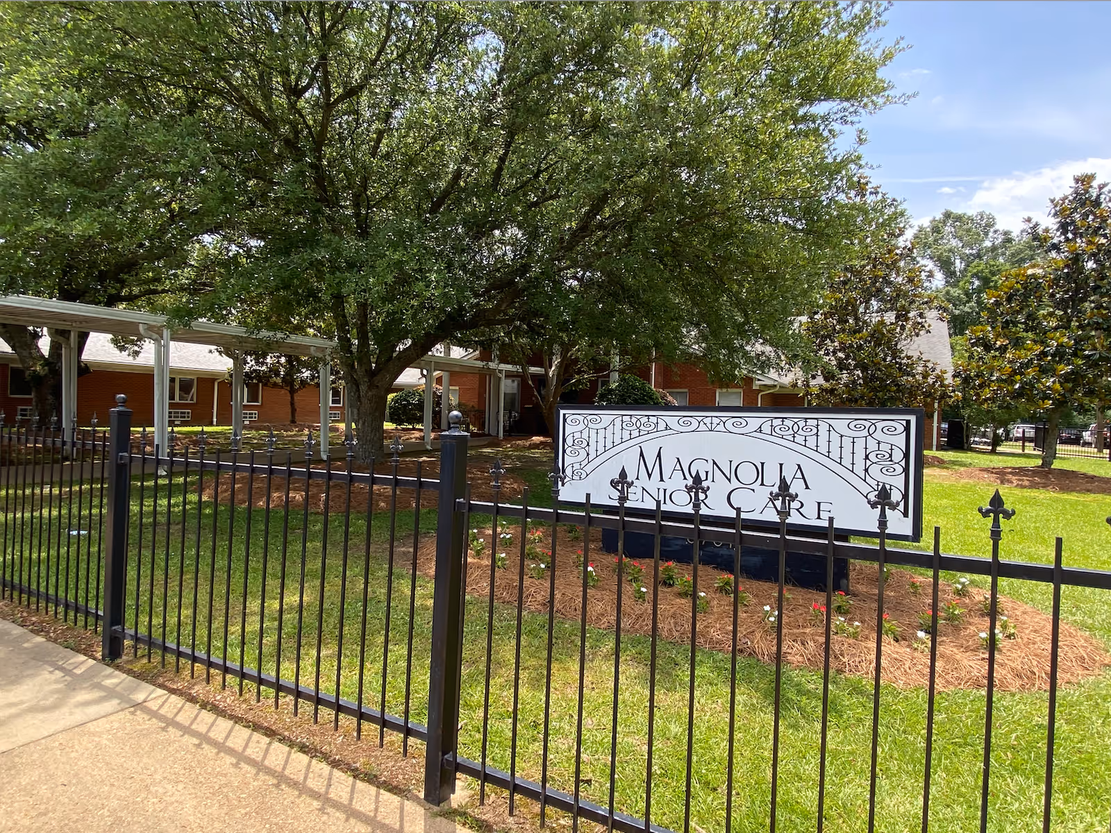 A Magnolia Senior Care sign on the lawn behind a black iron fence with trees and a brick building in the background.