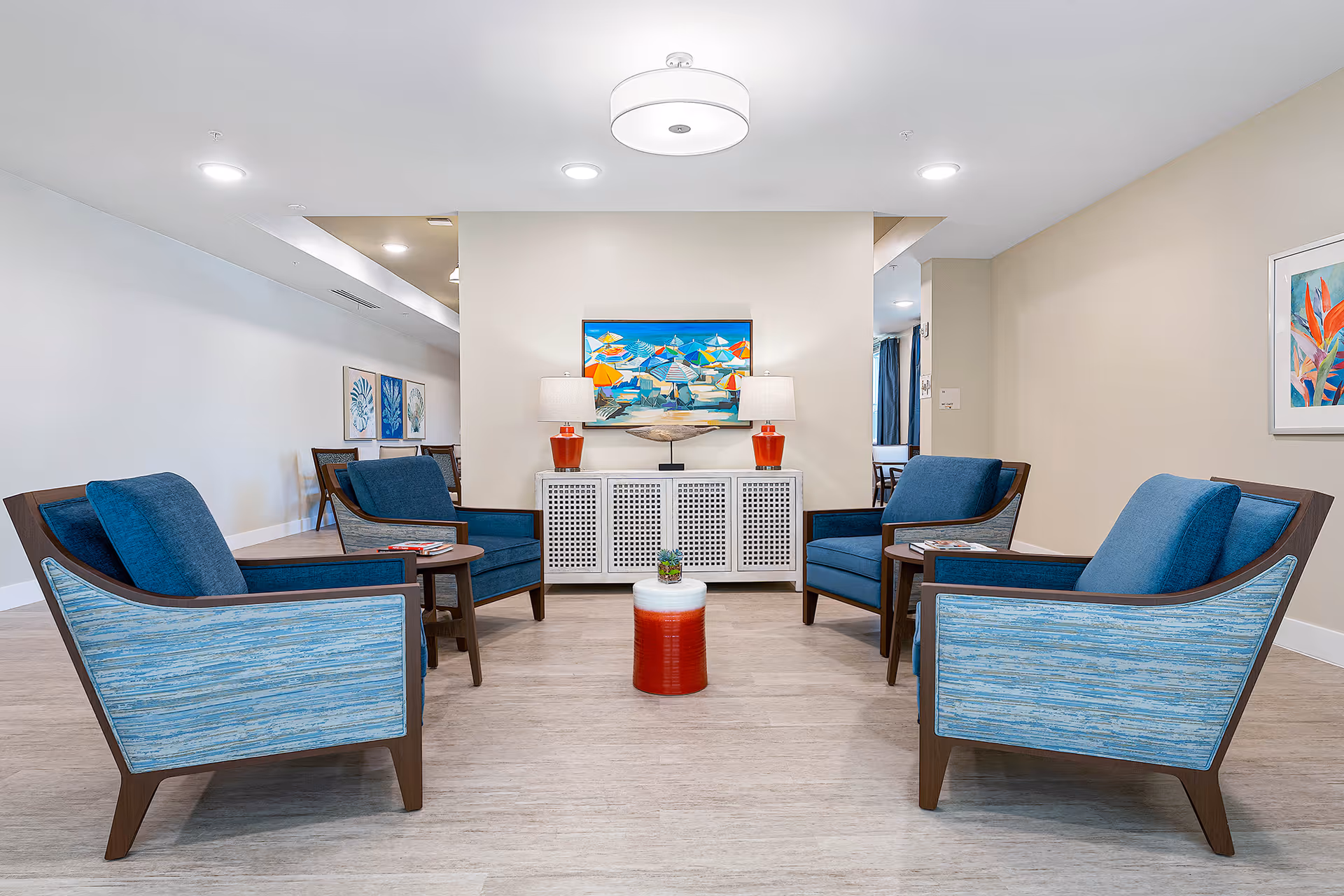 Bright common lounge with four blue armchairs arranged around small tables facing a cabinet topped with lamps and a colorful painting.