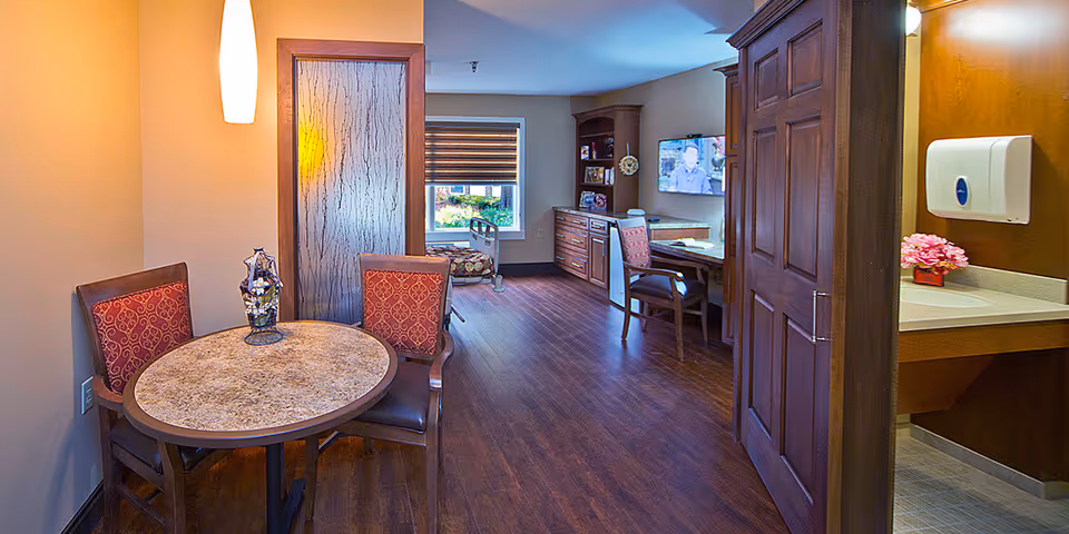 Interior senior living suite with a round table and chairs, wood floors, a desk and wall-mounted TV in the background, and a sink area visible to the right.