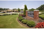 A landscaped outdoor area with a brick sign reading 'Harbor View Post Acute' surrounded by bushes and flowers, with a parking lot and building visible in the background under a partly cloudy sky.
