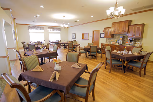 A spacious dining room with multiple tables covered in brown tablecloths, each set with folded napkins and small flower arrangements. The room has wooden flooring, several chairs around each table, large windows letting in natural light, and a kitchenette area with wooden cabinets and a countertop along one wall.