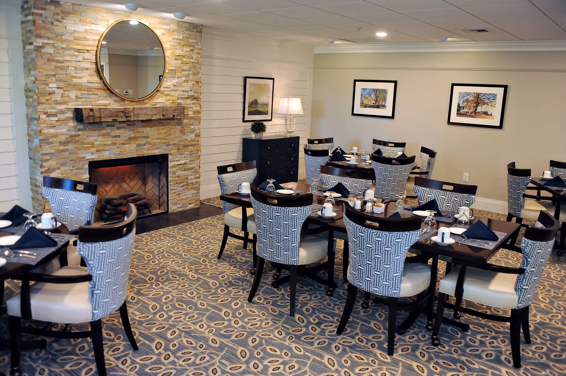 A cozy dining room with multiple tables set with cups, glasses, and napkins. The room features patterned chairs, a stone fireplace with a round mirror above it, framed artwork on the walls, and a patterned carpet.