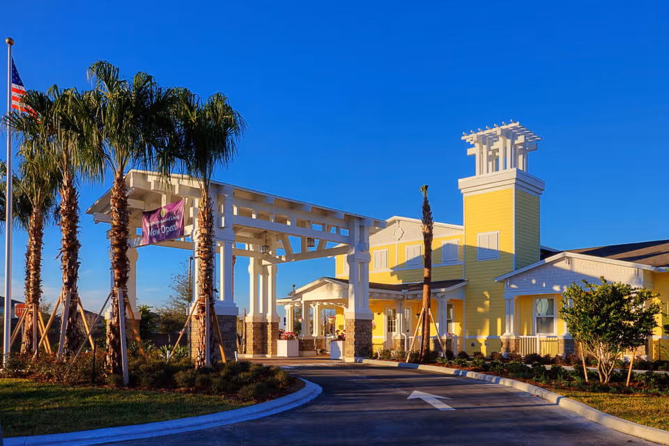 Exterior view of a senior living facility with a yellow building, white pergola entrance, palm trees, and an American flag. The sky is clear and blue, and there is a banner hanging on the pergola that says 'Now Open'.