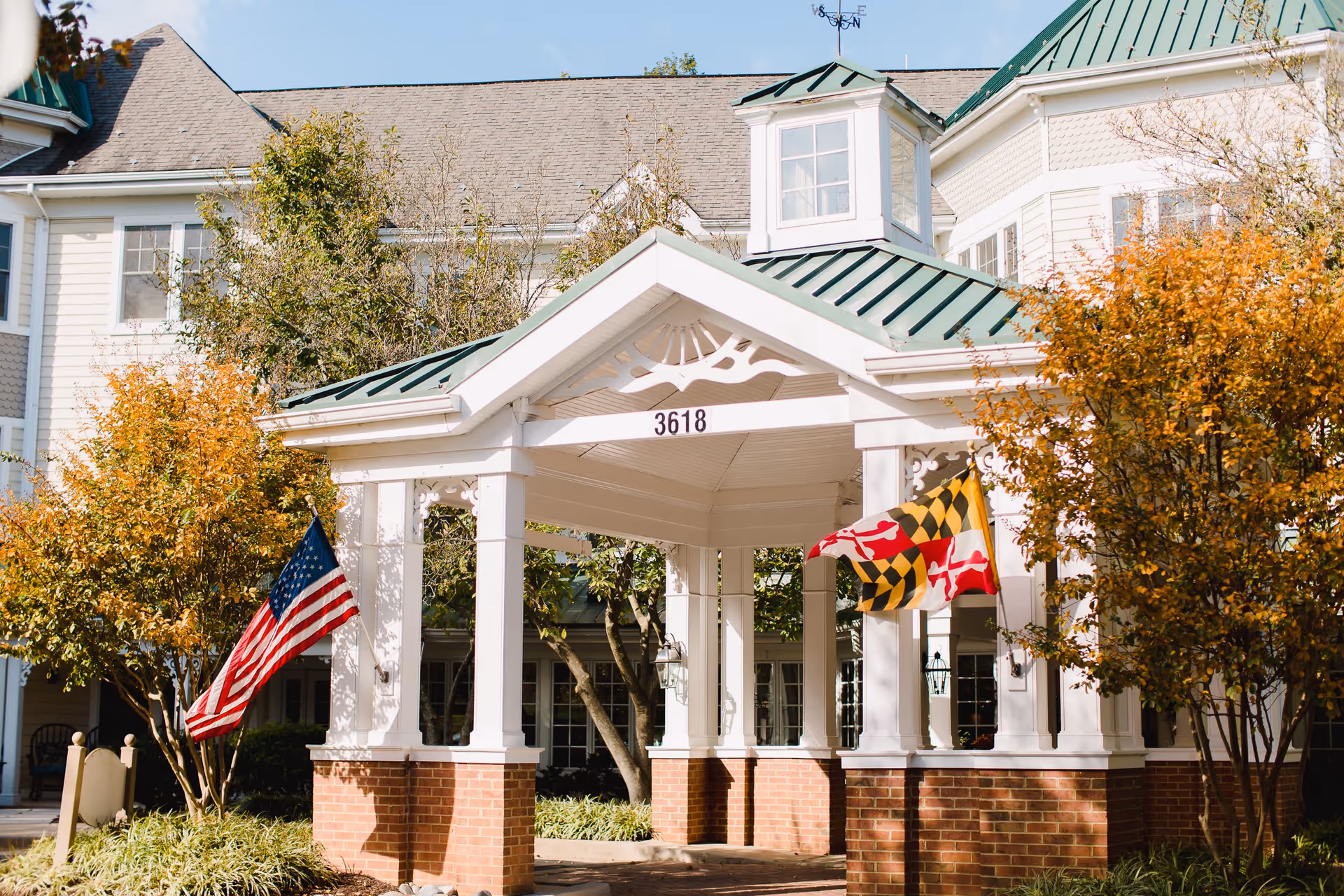 Entrance of a senior living facility with a white gazebo structure supported by brick pillars, displaying the number 3618. Two flags, the American flag and the Maryland state flag, are mounted on either side. The building behind has white siding, multiple windows, and a green metal roof. Trees with autumn foliage surround the entrance.