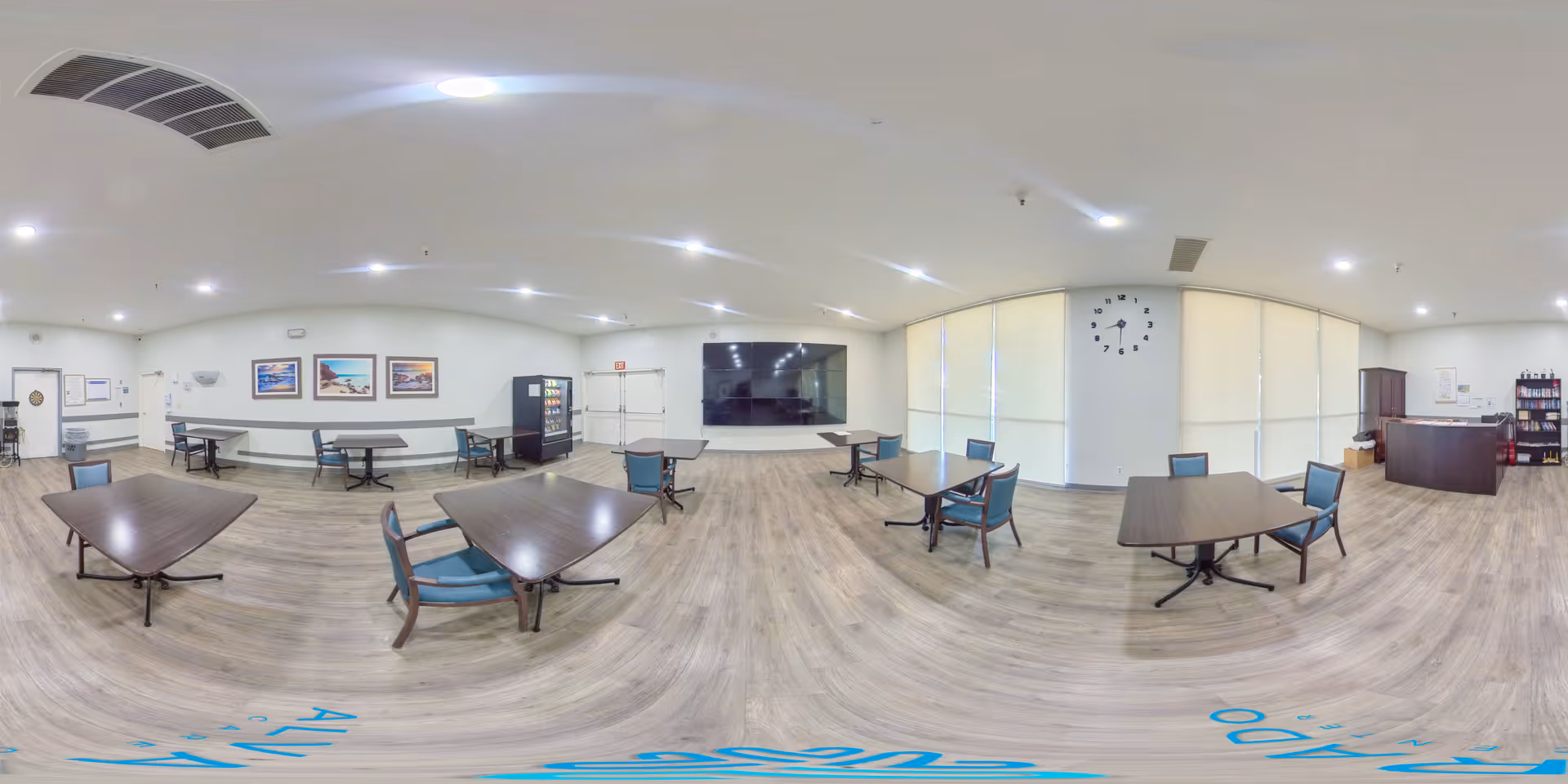 Spacious communal dining/activity room with several tables and chairs, a vending machine, wall clock and large windows.