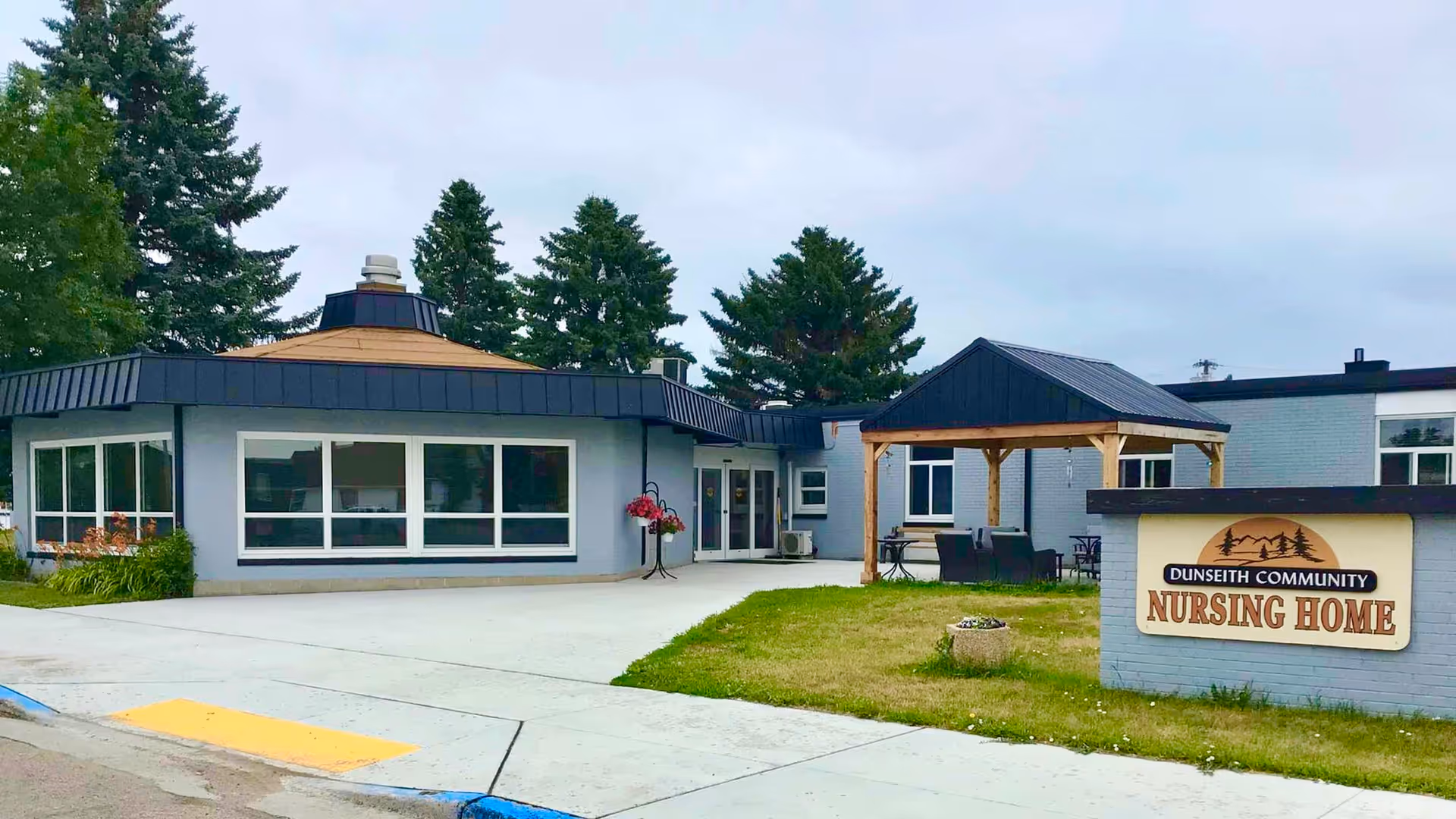 Exterior view of Dunseith Community Nursing Home showing a single-story building with large windows, a covered outdoor seating area with wooden posts, and a sign in front displaying the facility's name. There are trees in the background and a concrete walkway leading to the entrance.