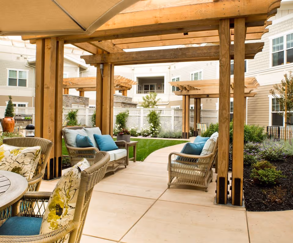 Outdoor seating area with wicker chairs and sofas featuring blue and floral cushions under wooden pergolas, surrounded by a garden with shrubs and a white lattice fence, adjacent to a multi-story building.