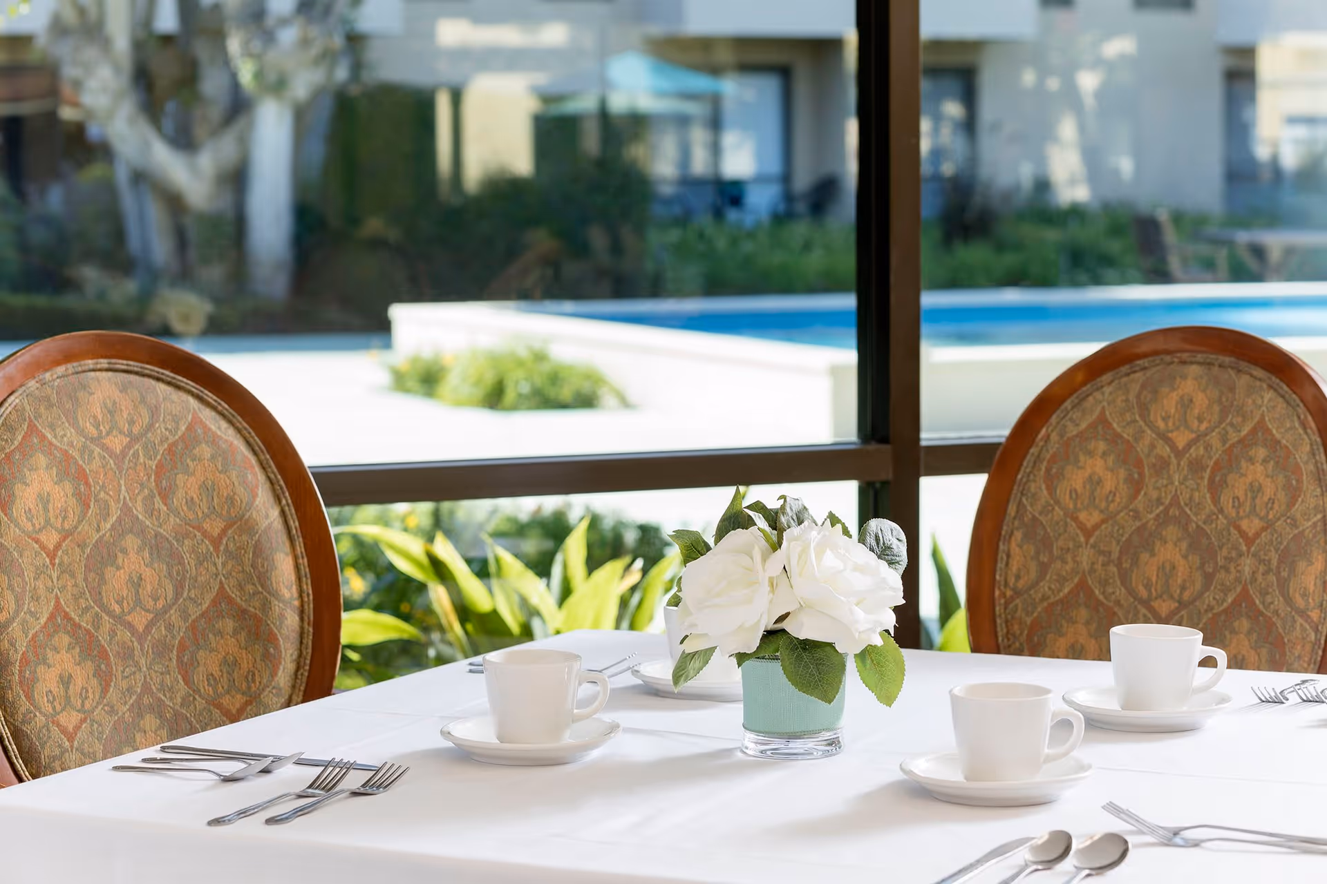 A dining table set with white cups, saucers, forks, and spoons, with a small vase of white flowers in the center. Two upholstered chairs with patterned fabric are positioned at the table. Large windows behind the table reveal a view of greenery and a swimming pool outside.