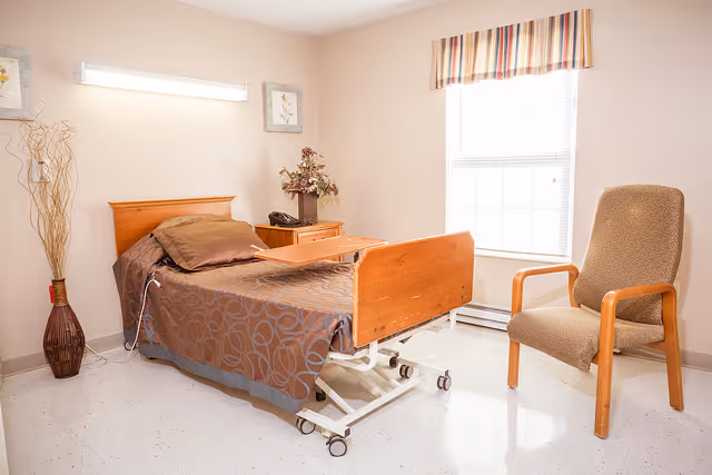 A well-lit room with a hospital-style bed covered with a brown patterned blanket and a matching pillow. The bed has a wooden headboard and footboard, and a wooden overbed table is positioned on top. To the right of the bed is a cushioned chair with wooden armrests. A window with blinds and a striped valance allows natural light into the room. On the left side, there is a tall vase with decorative branches and a small nightstand with a flower arrangement and a telephone. The walls are light-colored, and the floor is white with a glossy finish.