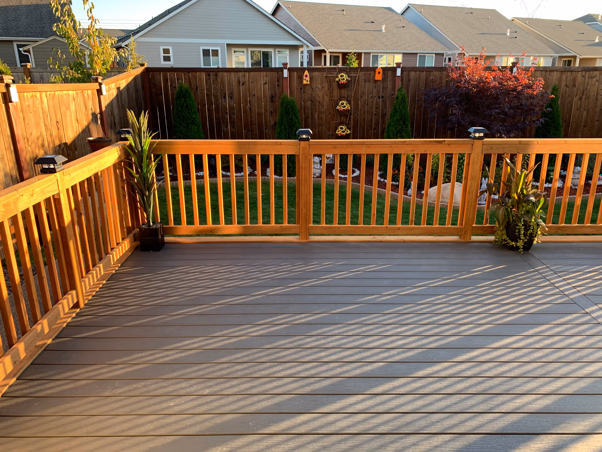 A wooden deck with railings overlooking a fenced backyard garden. The garden has green grass, small trees, decorative birdhouses, and potted plants on the deck. The scene is lit by warm sunlight.