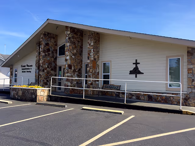 Exterior view of Curry Village Health and Rehabilitation of Cascadia building with stone pillars, a ramp with white railings, a bench, and a parking lot in front under a clear blue sky.