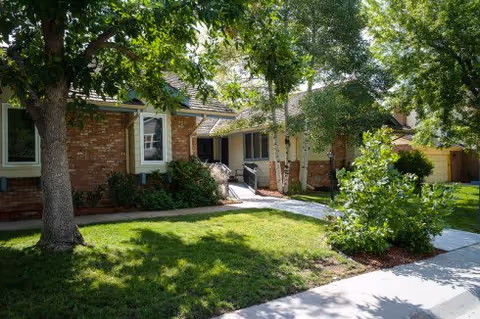 Exterior view of a single-story brick building with a sloped roof, surrounded by green grass, trees, and shrubs. A concrete walkway leads to the entrance, which has a small ramp and handrails.