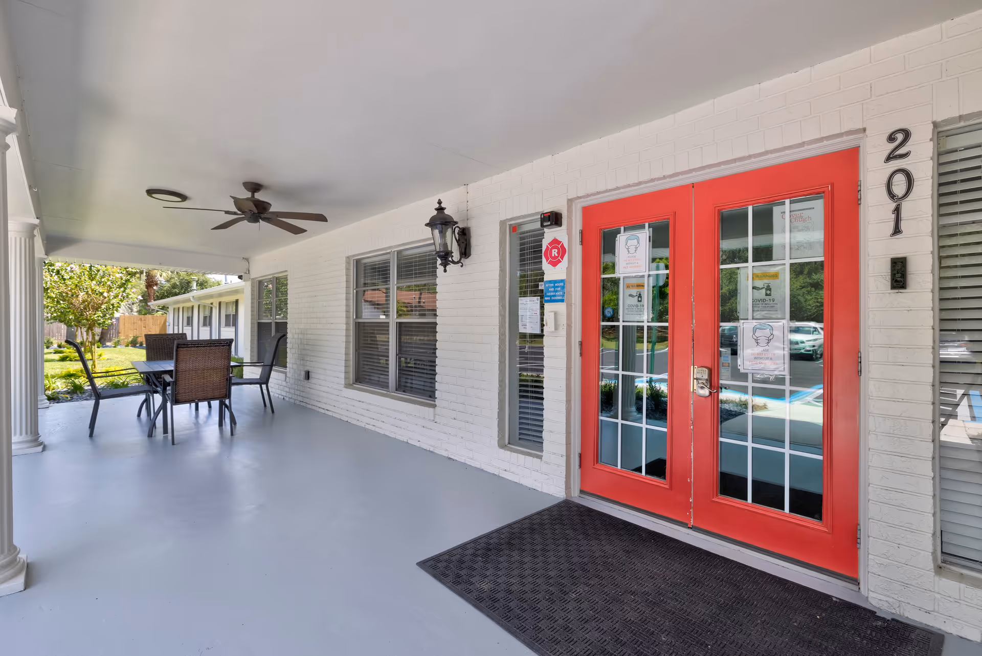 Covered outdoor patio area with a ceiling fan, a table, and four chairs. The patio has white brick walls and a bright red double door entrance with glass panels. The door has several COVID-19 related signs posted on it. The building number 201 is displayed next to the door. There is a black outdoor wall lantern light fixture mounted on the wall near the door. Green grass and trees are visible in the background.