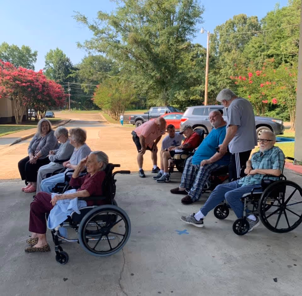 A group of elderly people, some in wheelchairs, sitting and socializing outside on a concrete area near a parking lot with trees and flowering bushes in the background. One man is bending down to talk to a seated person, while others are seated or standing nearby.