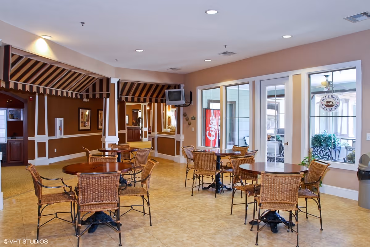 A spacious dining area with several round wooden tables and wicker chairs. The room has beige walls and tiled floors, with large windows and glass doors leading to an outdoor patio area. There is a small wall-mounted TV and a Coca-Cola vending machine visible through the windows.