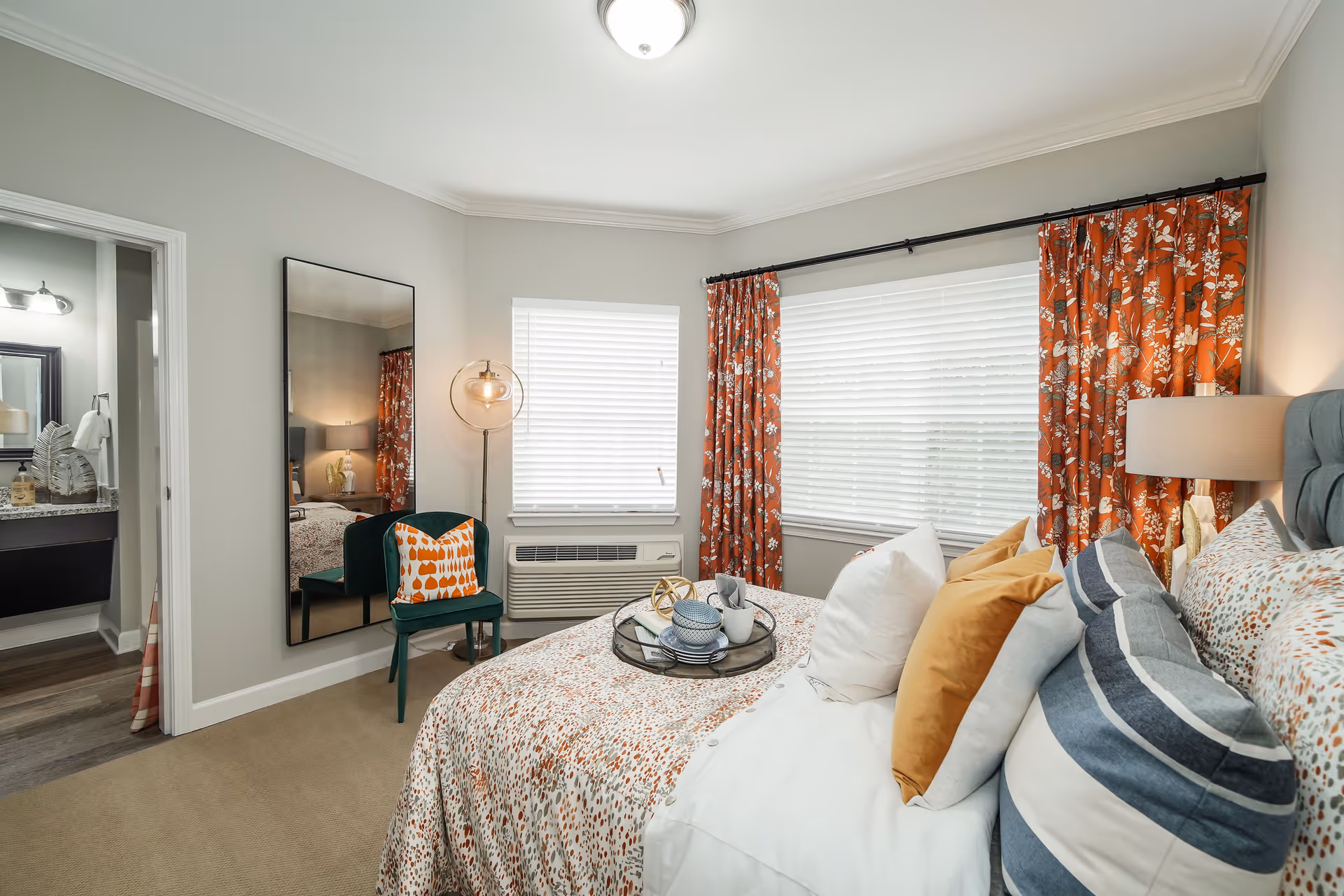 A cozy bedroom with a bed adorned with multiple pillows in white, mustard yellow, and blue striped patterns. The bedspread has a subtle spotted design. Next to the bed is a lamp on a nightstand. The room features two windows with white blinds and orange floral curtains. A green chair with an orange and white patterned pillow sits beside a full-length mirror mounted on the wall. A bathroom is visible through an open door, showing a sink and mirror.