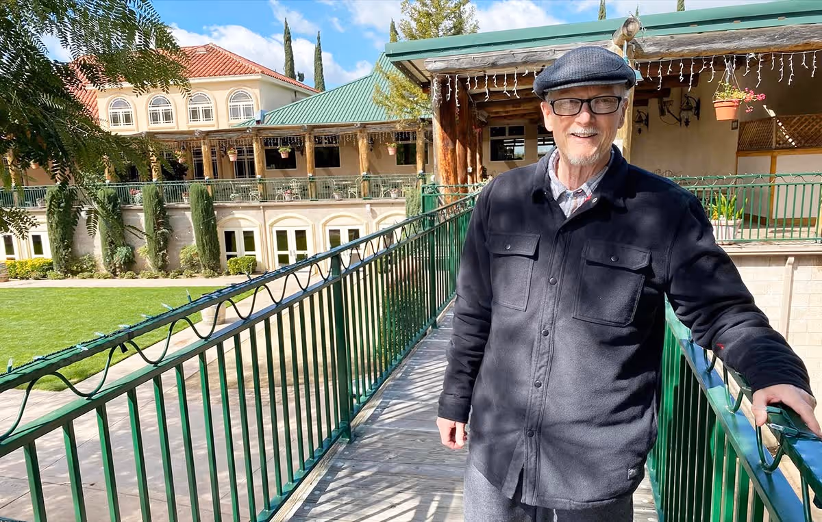 An elderly man wearing a black jacket, gray pants, glasses, and a flat cap stands on a green metal-railed walkway outside a building with a green roof and beige walls. The background shows a well-maintained lawn, trees, and a multi-story building with arched windows under a partly cloudy sky.