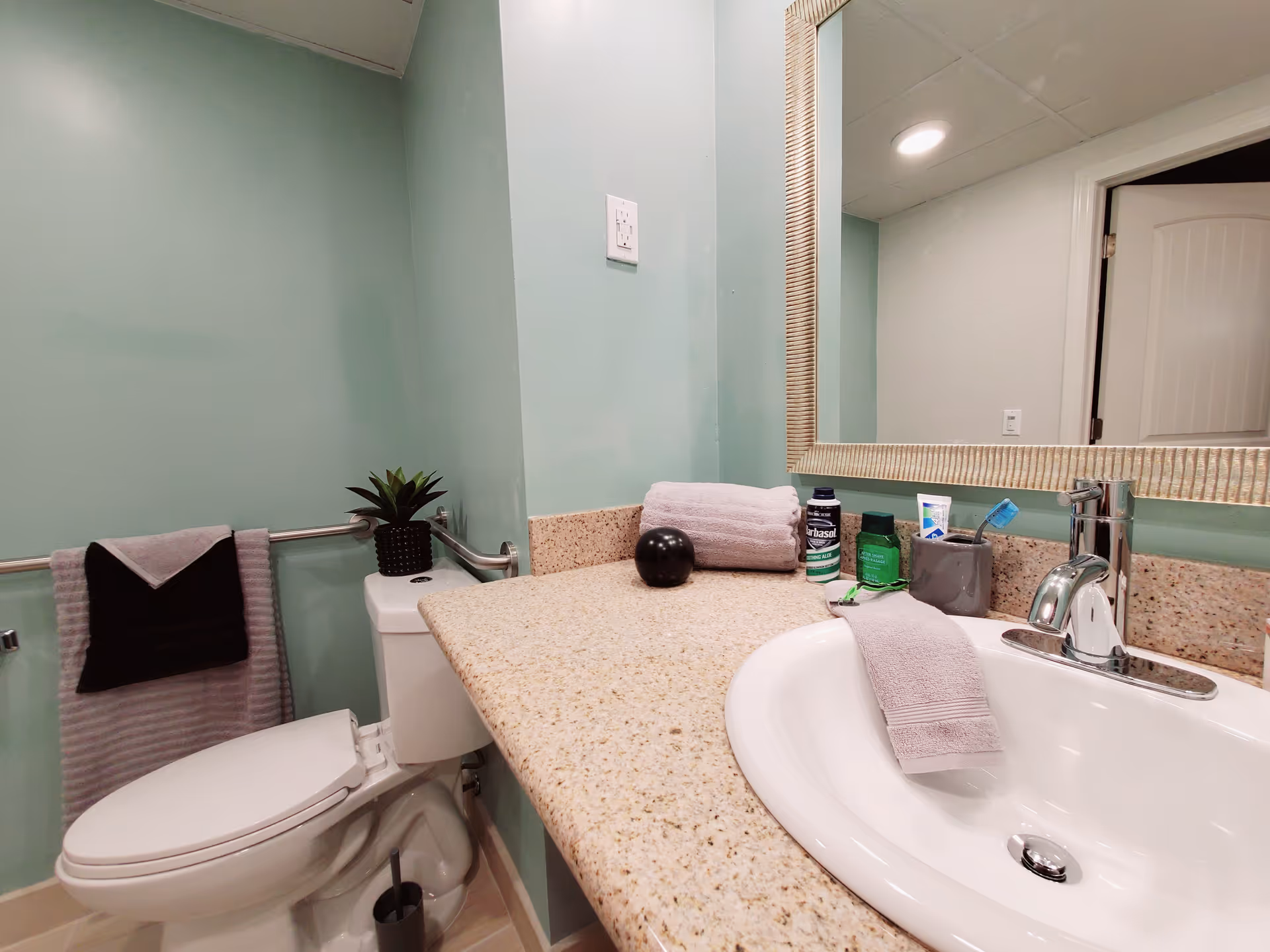 Bathroom with mint-green walls showing a sink set in a speckled countertop, a toilet, large mirror, towels, and toiletries.