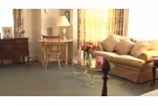 A cozy living room with a beige sofa adorned with cushions, a glass coffee table with a flower vase and a card, a wooden desk with a chair and a lamp, and a wooden dresser with framed photos. The room is softly lit by natural light coming through yellow curtains.